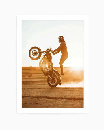 "Framed photograph of a person performing a motorcycle wheelie at sunset, printed on fine art paper and displayed in a white frame with white border. The composition features a person in a helmet and dark clothing standing on a motorcycle, with the front wheel lifted high in the air and the back wheel kicking up dust. The setting sun creates a warm, golden glow across the sandy ground and bright sky."
