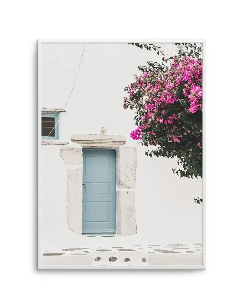 Framed photography art print of a white stucco building with a light blue door and window, surrounded by vibrant pink bougainvillea flowers. The composition captures the bright sunlight and traditional Greek island architecture, with a white frame and white border.