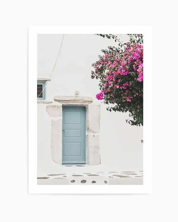 "Photography art print featuring a white-washed building with a light blue door and window, framed by a vibrant pink bougainvillea plant on the right. The ground in front of the door is a white cobblestone path with dark grey, irregular-shaped stones."