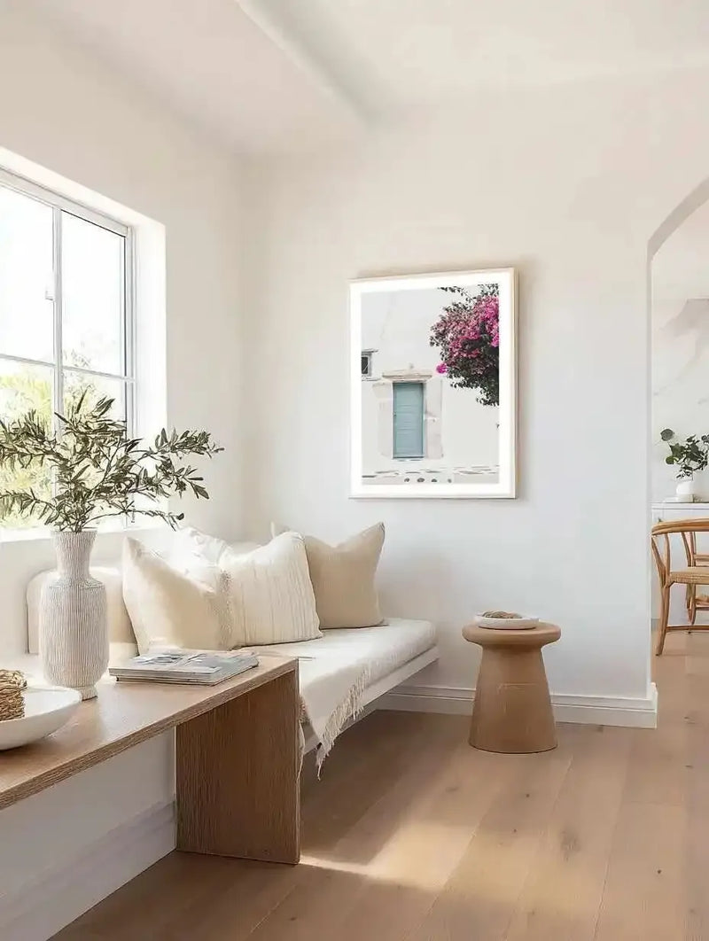 Lifestyle shot of a minimalist living room with a framed art print displayed above a white daybed. The art print features a photography of a white building with a light blue door and a vibrant pink bougainvillea tree on the right side. The room includes a wooden bench, a white vase with green foliage, cream-coloured pillows, and a light brown wooden stool.