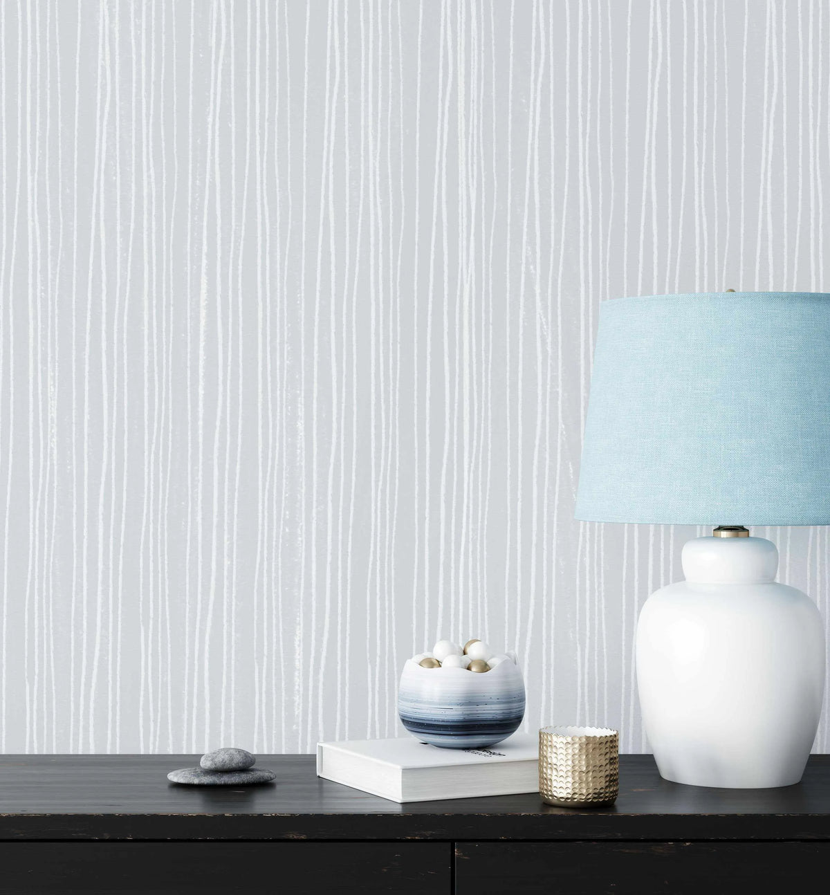 Lifestyle shot featuring a wall covered in Subtle Stripes Wallpaper in Blue, a light grey wallpaper with a repeating pattern of thin, hand-drawn vertical white lines of varying thickness and slight waviness, creating a subtle, organic stripe effect. The wallpaper is visible behind a dark wooden console table with a white ceramic lamp with a light blue fabric shade, a small blue and white striped bowl with white and gold decorative balls, a white book, a gold textured votive, and two stacked grey stones.