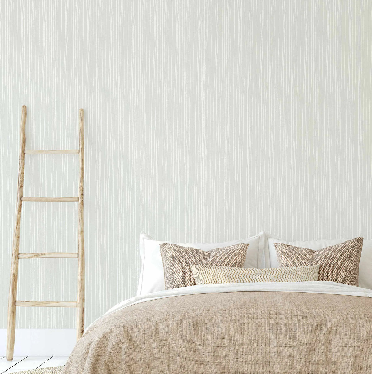 Lifestyle shot of a bedroom featuring Subtle Stripes Wallpaper in Sage. The wallpaper has a repeating pattern of thin, hand-drawn vertical lines in white on a soft sage green background, creating a subtle, organic texture. The wallpaper covers the wall behind a bed with a light brown textured duvet and pillows. A rustic wooden ladder leans against the wall on the left side of the frame.