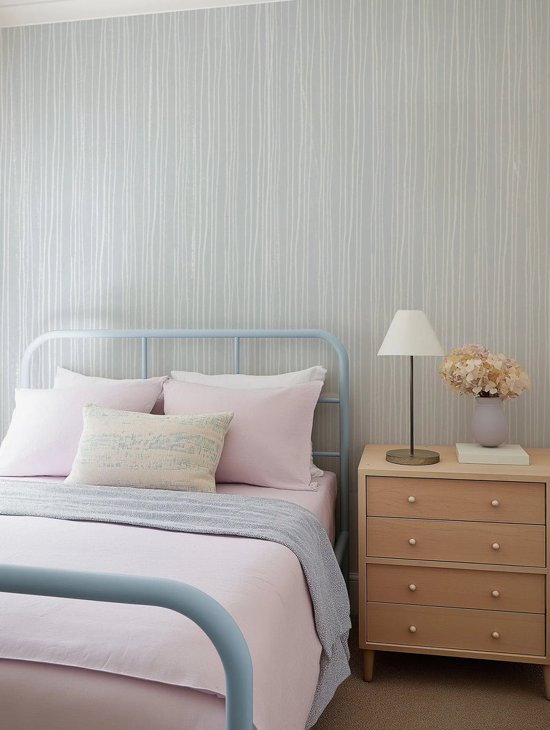 Lifestyle shot of a bedroom featuring a light blue wallpaper with a subtle, repeating pattern of thin, vertical white stripes with a hand-drawn, organic quality. The wallpaper covers the wall behind a light blue metal bed frame with pink bedding and a textured grey throw blanket. A light wood bedside table with a lamp and a vase of dried flowers sits to the right of the bed.