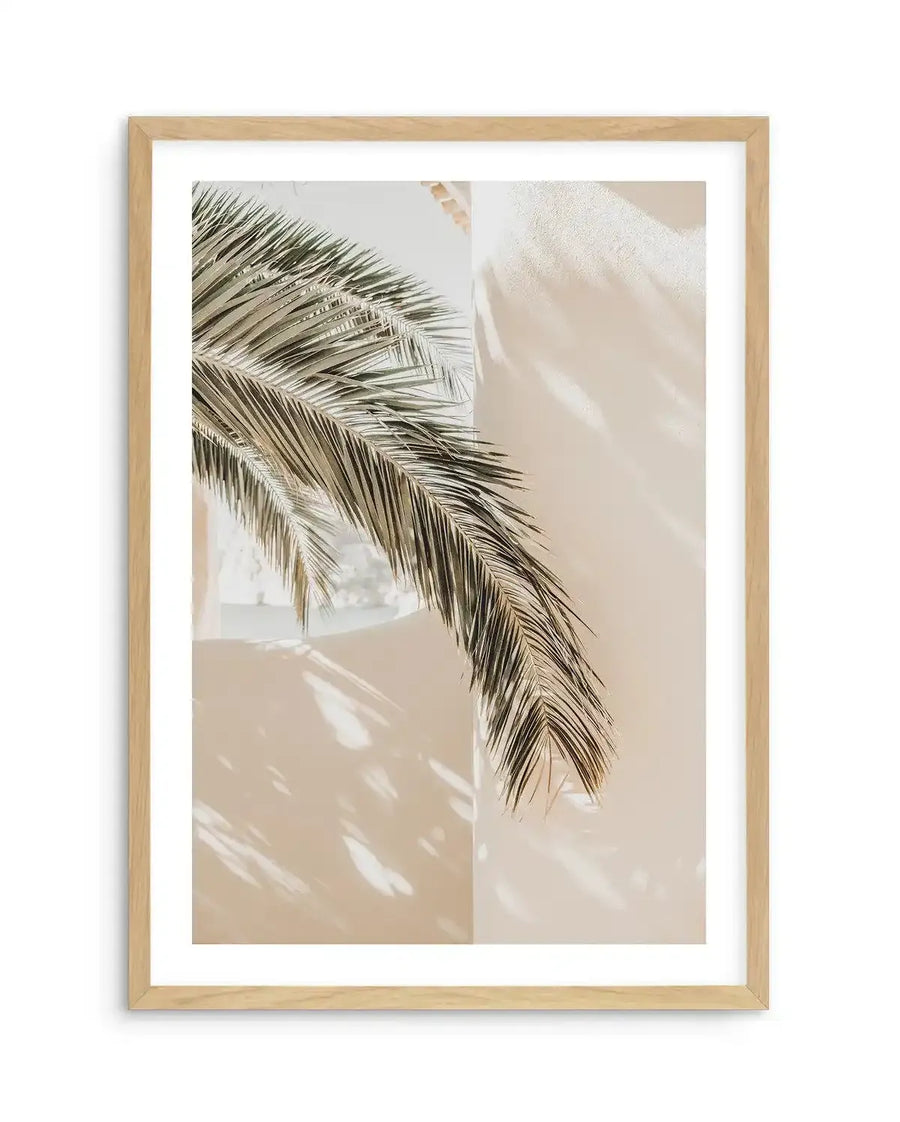 Framed photography art print of a palm frond casting shadows on a cream-colored wall, with a natural oak frame and white border. The palm frond is in the upper left, extending diagonally downwards to the right, with its dark green leaves creating intricate shadow patterns on the sunlit wall.