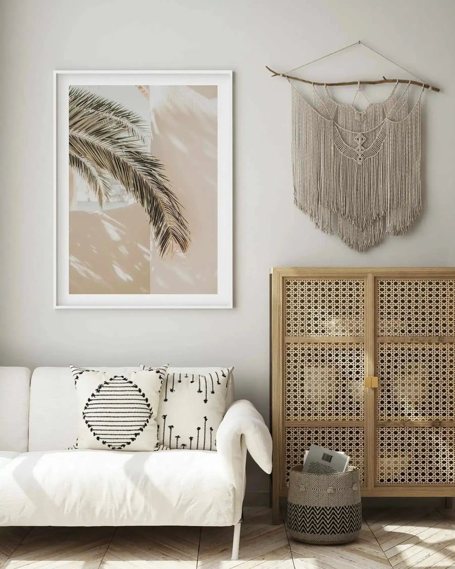 Large framed art print displayed above a white sofa in a minimalist living room. The print features a close-up photograph of a palm frond in natural green and brown tones, casting shadows on a light beige wall, with a white frame and white border. The room includes a macrame wall hanging, a rattan cabinet, and a woven basket.