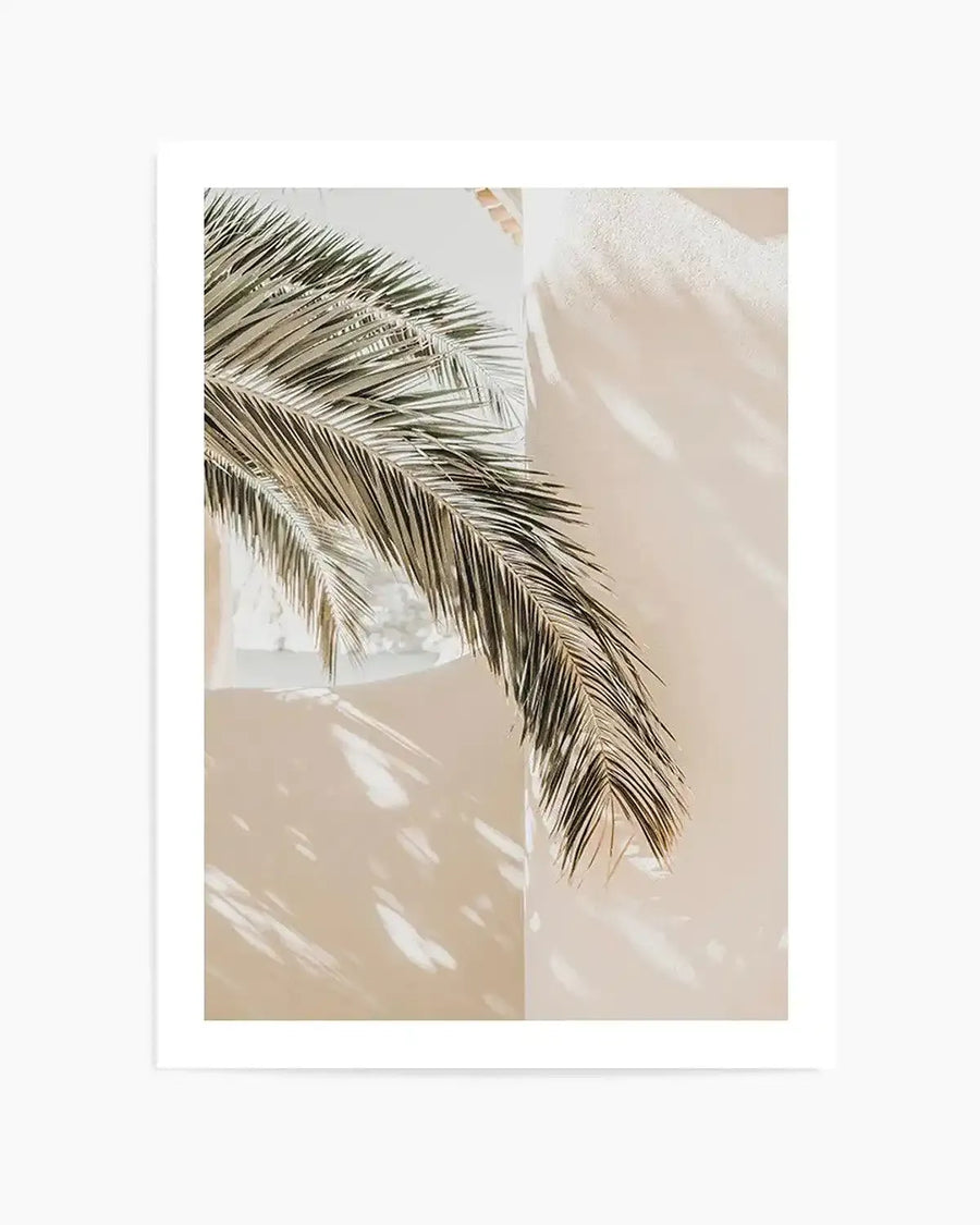 Framed photography art print capturing a sun-drenched scene with a palm frond against a cream-colored wall. The palm frond, in shades of muted green and brown, extends diagonally from the upper left to the lower right, casting dappled shadows on the textured wall. The bright sunlight creates a warm, airy mood.