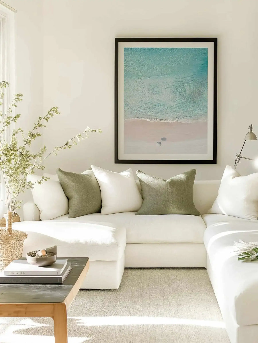 Lifestyle shot of a minimalist living room featuring a large framed art print displayed above a white sectional sofa. The print is an aerial photograph of a beach with a pale pink sandy shore meeting shallow turquoise water, with two small dark figures on the sand. It is displayed in a black frame with a white border. The sofa is adorned with white and sage green throw pillows. A light wood coffee table with books and a decorative bowl sits in front of the sofa.