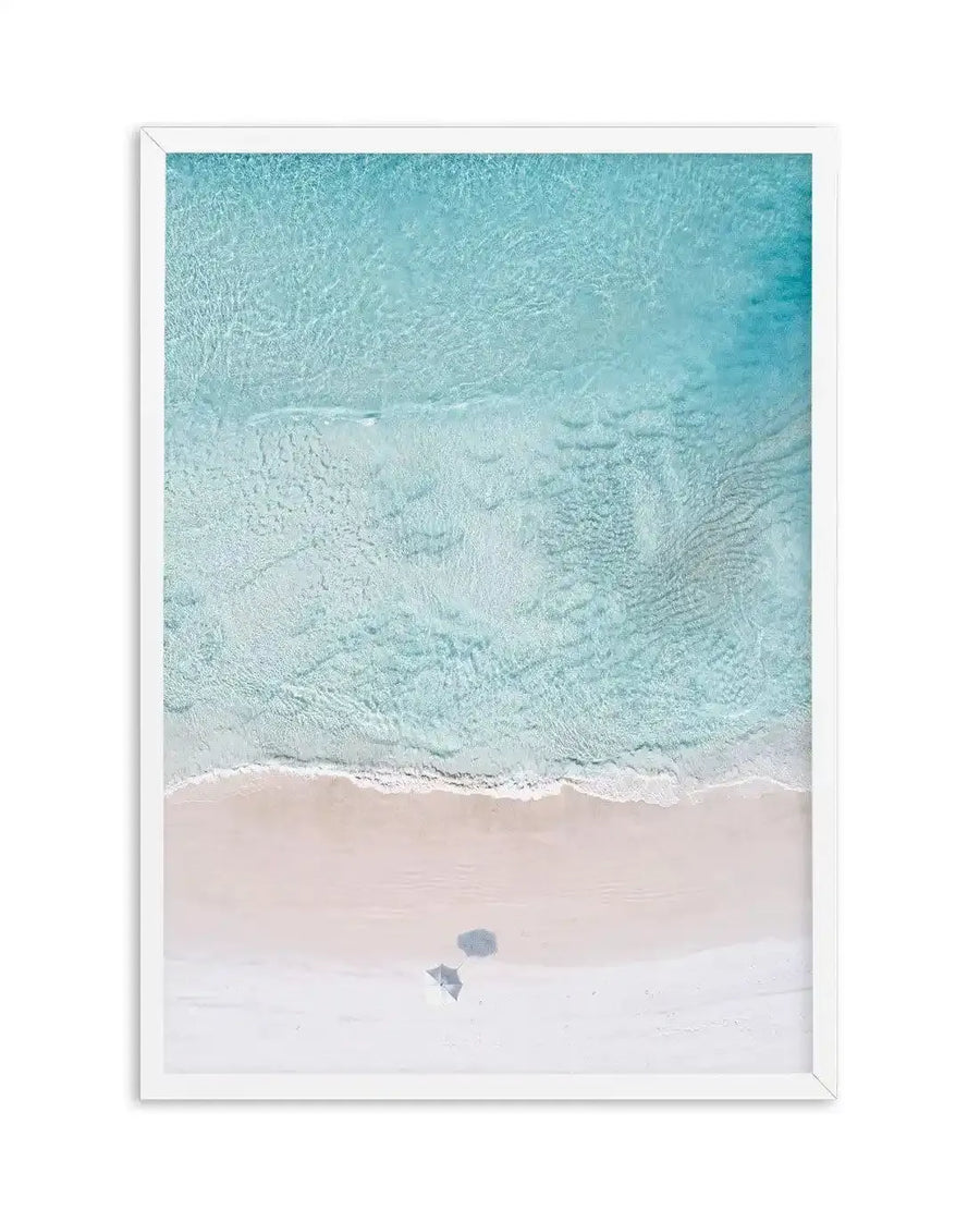 Framed aerial photograph of a pristine beach with turquoise ocean and white sand, displayed in a white frame with white border. The composition captures gentle waves breaking on the shore, with two small shadows on the sand, suggesting beach umbrellas or figures.