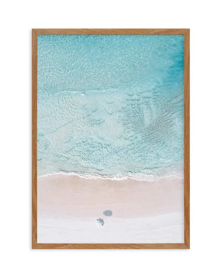Framed aerial photograph of a pristine beach with clear turquoise water and white sand, displayed in a natural oak frame with white border. The composition shows gentle ripples on the water and a soft wave breaking on the shore, with two small shadows visible on the sand.