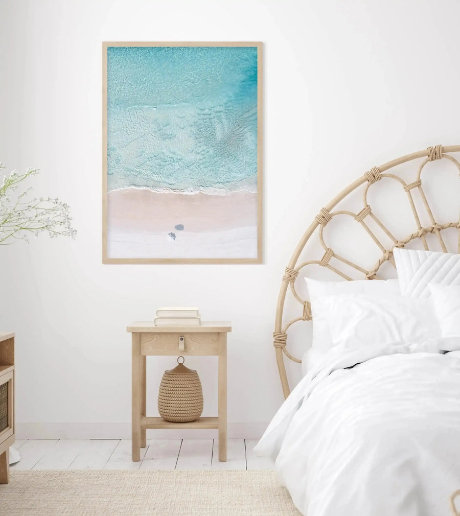 Lifestyle shot of a minimalist bedroom featuring a framed aerial photograph of a beach. The print, displayed above a light wood bedside table, captures a pale pink sandy beach with gentle turquoise waves breaking on the shore, and two small beach umbrellas. The room has a white wall, a light wood rattan headboard, white bedding, and a textured cream rug.
