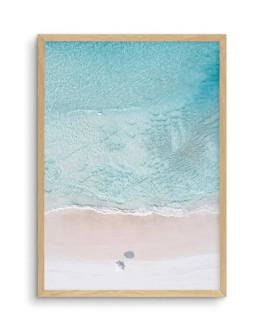 Framed aerial photograph of a beach scene with natural wood frame and white border. The composition features clear turquoise ocean water with gentle ripples, meeting a stretch of light pink sand where a small white umbrella and a grey beach towel are visible.