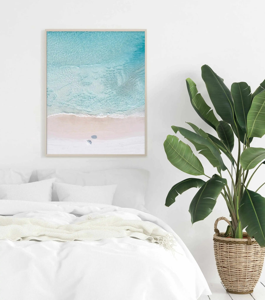 Lifestyle shot of a minimalist bedroom featuring a framed aerial photograph of a beach scene displayed above a bed with white bedding. The print shows clear turquoise ocean water meeting a soft pink sandy beach, with gentle waves breaking at the shore and two small figures with an umbrella on the sand. The print has a natural wood frame and white border. A large potted plant with broad green leaves sits in a woven basket beside the bed.