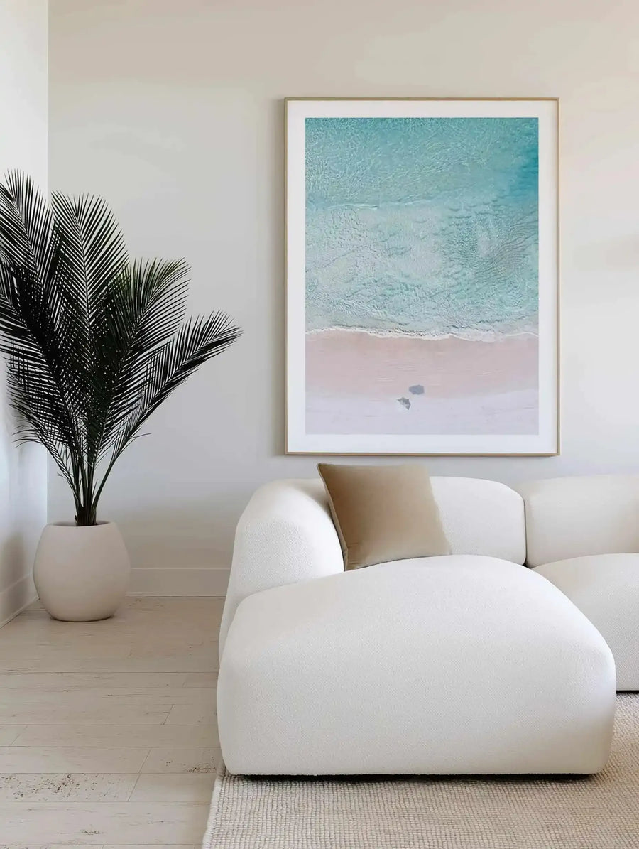 Large framed art print displayed above a curved white sofa in a minimalist living room. The print is an aerial photograph of a beach with a wide expanse of turquoise ocean meeting a pale pink sandy beach, with two small dark shapes on the sand, with a natural wood frame and white border. The room includes a large green palm plant in a white pot and a light brown cushion on the sofa.
