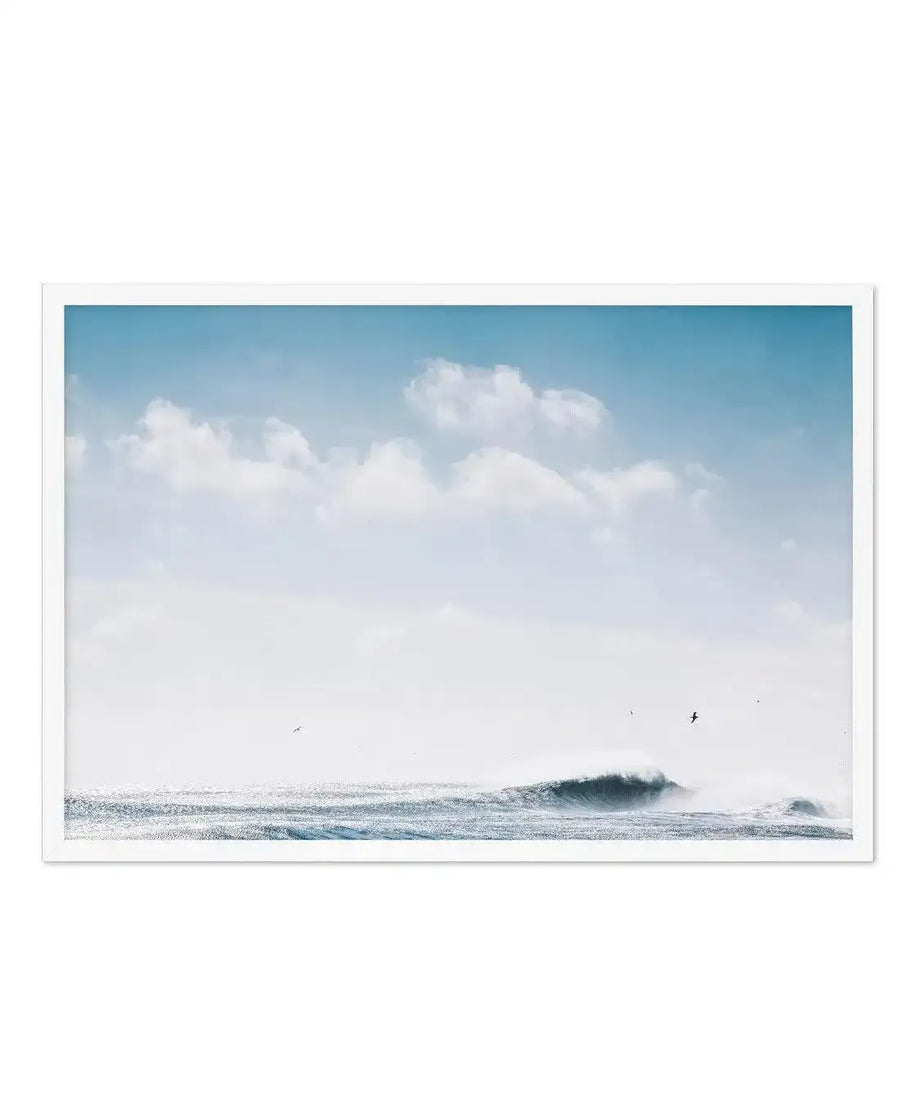 Framed photography art print featuring a serene ocean scene with a large wave breaking on the right, under a bright blue sky with scattered white clouds. The water glistens with sunlight, and a few birds are visible in the distance, with a white frame and white border.