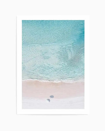 Aerial photography art print of a tranquil beach scene with clear turquoise water meeting a pale pink sandy shore. Gentle white waves lap at the edge of the sand, and two small, dark shadows are cast on the beach, suggesting people or objects. The water shows subtle ripples and variations in depth, creating a serene, natural composition.