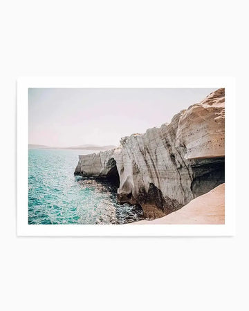 "Framed photograph of a coastal scene in Greece with white rock cliffs and turquoise water, with white frame and white border. The composition features white rock formations with natural caves and arches, with gentle waves lapping at the shore under a bright, clear sky."