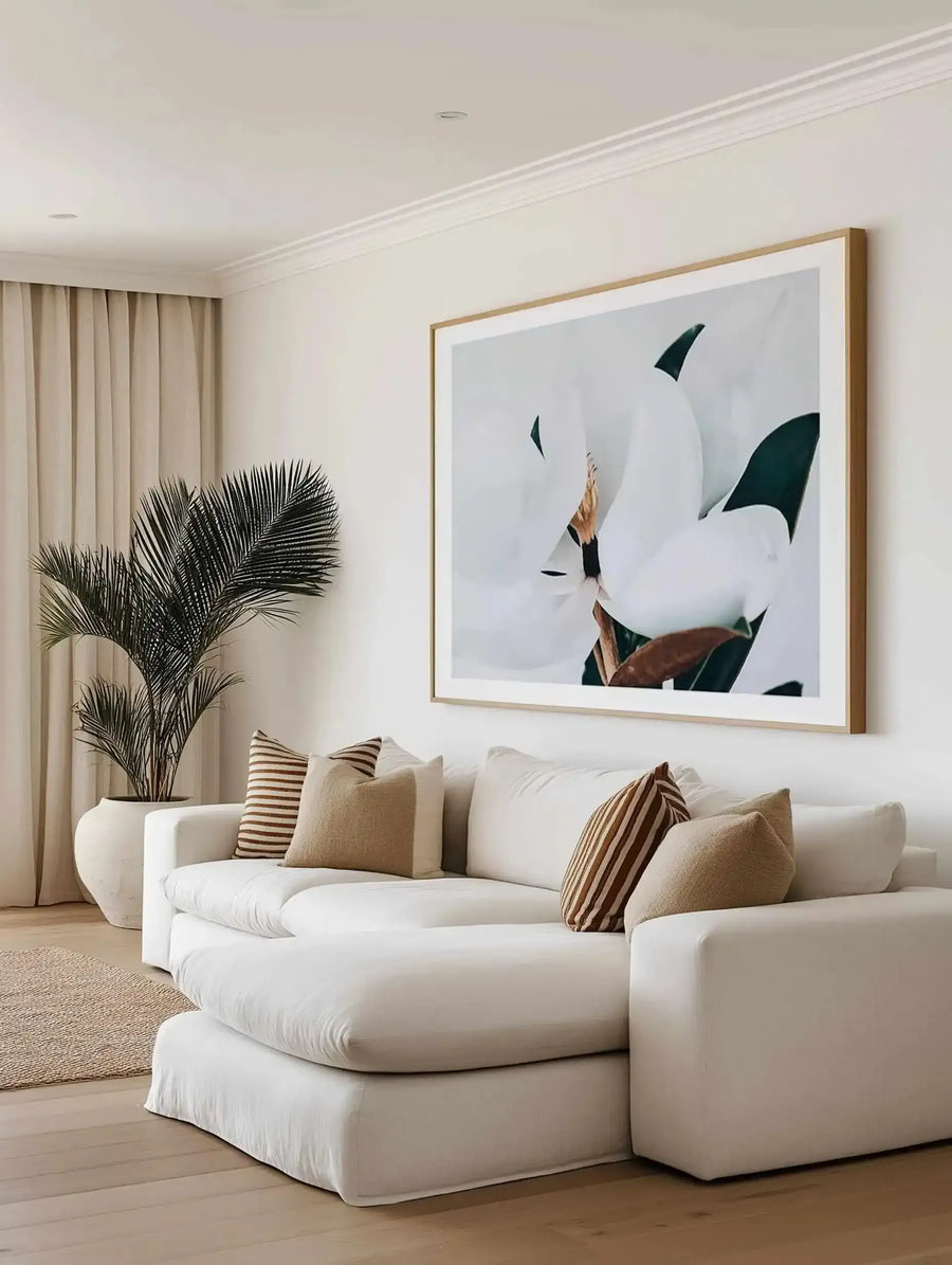 Summer Magnolia art print displayed in a natural wood frame with white border, hung above a curved cream upholstered sofa in a minimalist living room. The artwork features a person in dark clothing with arm raised, surrounded by dark green and teal leaf shapes against a soft gray-white background, creating a serene, botanical aesthetic. The room includes a potted black palm plant, striped and solid tan throw pillows, beige curtains, neutral area rug, and light wood flooring.