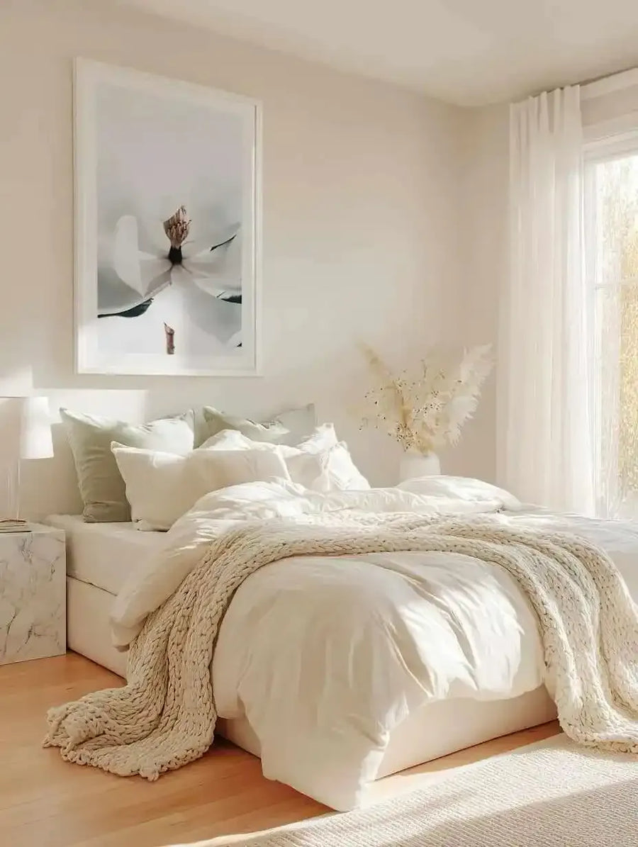 Large framed art print displayed above a bed in a minimalist bedroom. The print features a close-up photograph of a white magnolia flower with a brown and orange center, against a soft white background, with a white frame and white border. The room includes a bed with white bedding, sage green and white pillows, a chunky knit cream throw, a white marble bedside table with a lamp, and a vase of dried pampas grass.
