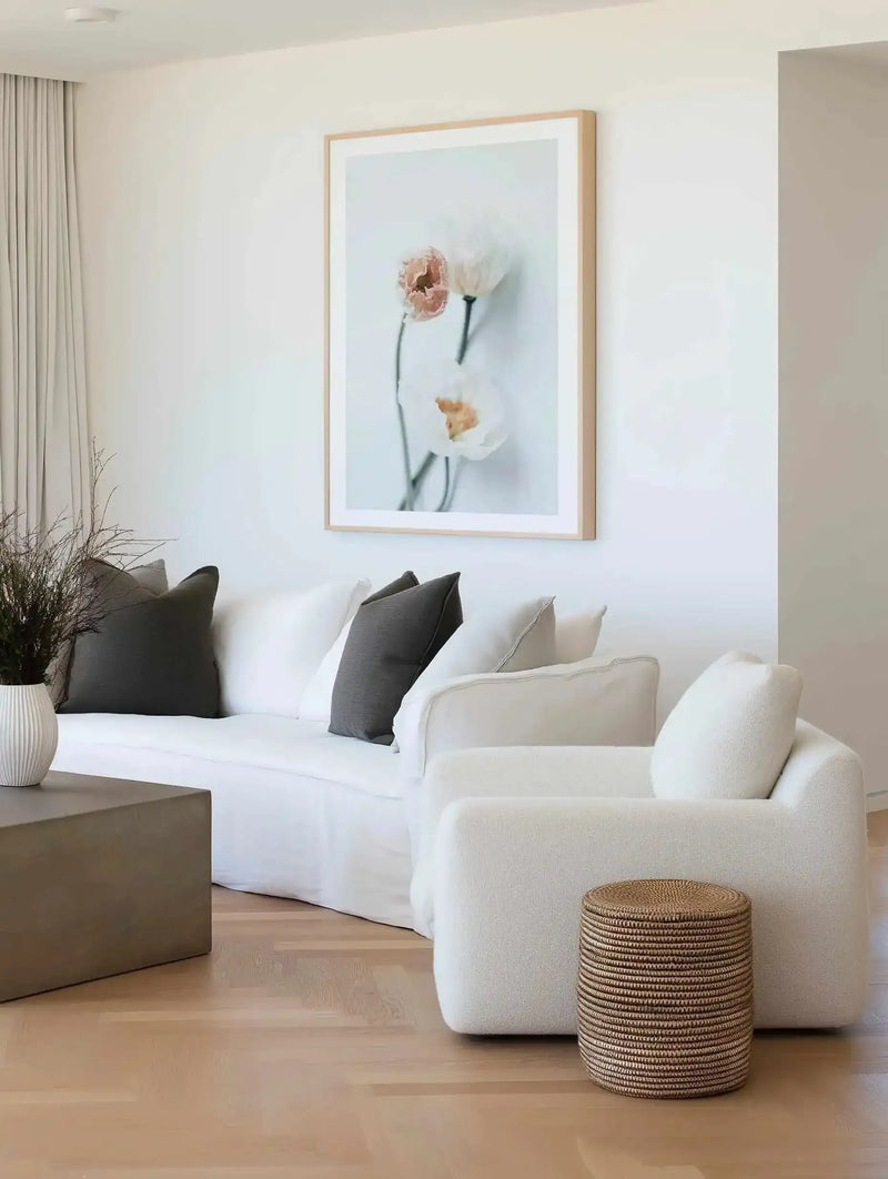 Summer Poppies I art print displayed in a light wood frame with white border, hanging on a white wall above a cream curved sofa. The artwork features two delicate poppy flowers in soft peachy-pink and cream tones with dark green stems against a pale blue-grey background, painted in a loose, watercolor style. The print is shown in a minimalist living room setting with natural wood flooring, grey and black accent pillows, a woven side table, and neutral décor.