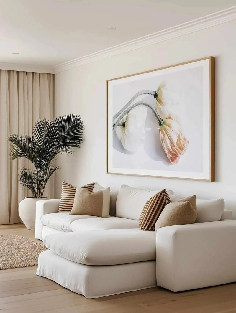 Framed art print titled Summer Poppies II displayed on a cream wall above a curved white sofa in a minimalist living room. The watercolor illustration features delicate poppy flowers in soft peachy-coral and cream tones with gray-green stems, rendered in a botanical style against a light background. The print has a natural wood frame with white border. The room includes a potted black palm plant, striped throw pillows in tan and brown, and beige curtains, creating a warm, neutral aesthetic.