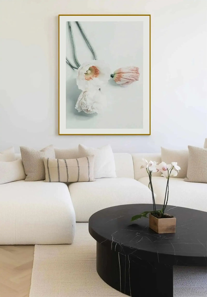 Framed art print featuring two delicate poppy flowers with blush pink petals and cream-colored ruffled blooms, rendered in soft watercolor style against a pale gray-green background. Gold-toned frame with white border displayed above a cream linen sectional sofa with neutral pillows in a modern living room setting.