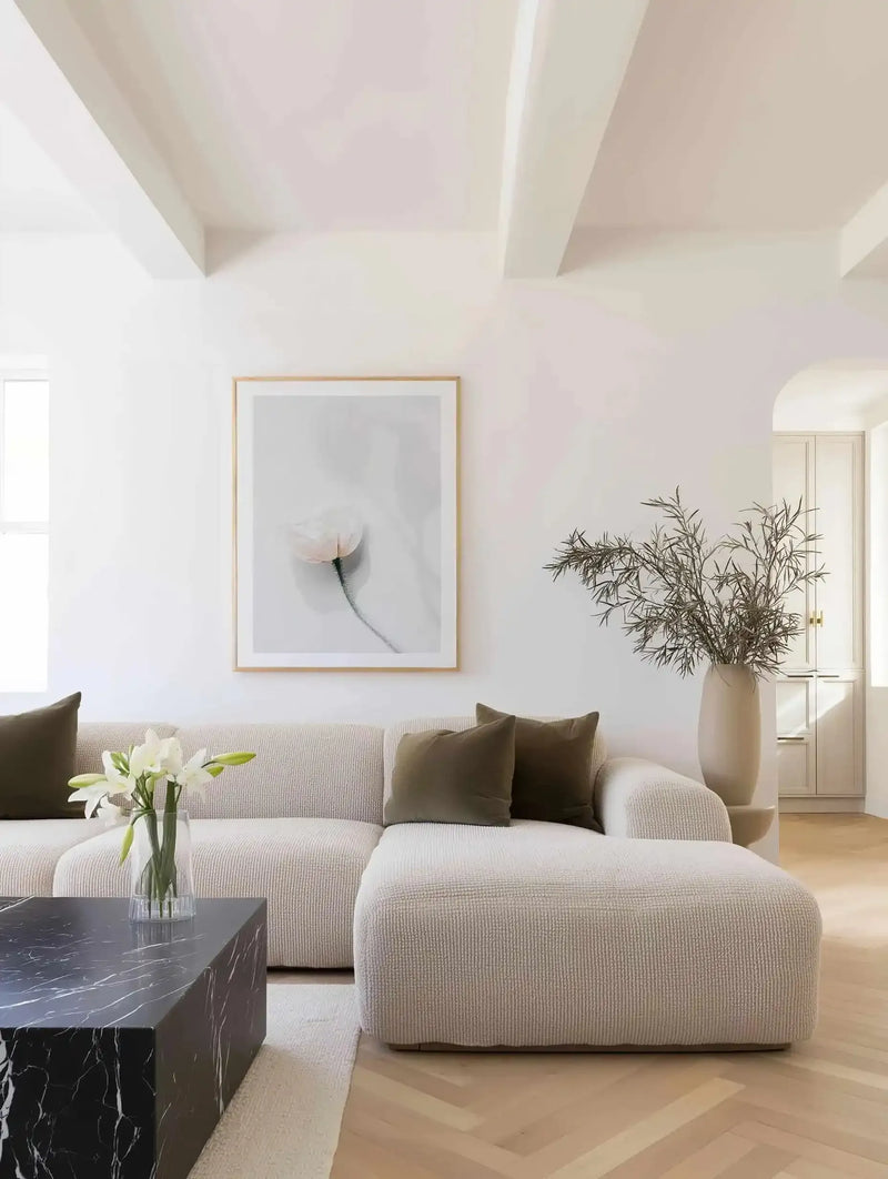 Large framed art print displayed above a cream textured sectional sofa in a bright, minimalist living room. The print features a single, delicate light pink poppy with a long, thin stem against a soft grey background, with a natural wood frame and white border. The room includes dark olive green throw pillows, a black marble coffee table, and a tall cream vase with dried branches.