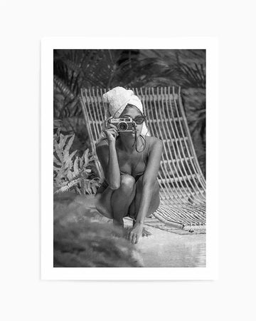 Black and white art print titled "Summer of Love II" by Mario Stefanelli, featuring a vintage photograph of a woman in a bikini crouching by water. She wears sunglasses and a white head wrap, holding a vintage camera up to her face. Palm fronds and a striped beach chair are visible in the blurred background. The photograph has a grainy, mid-century quality with rich tonal contrast. The print is mounted on a white border and framed simply, presented as a vertical composition.