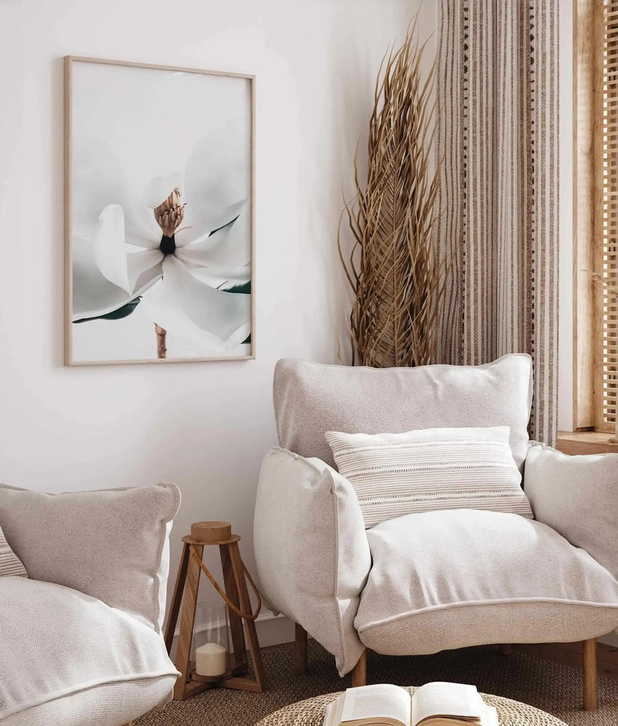 Summer Magnolia art print displayed in a light wood frame with white border, hung on a white wall above a cream linen armchair. The artwork features a close-up botanical photograph of a white magnolia flower with delicate petals and dark green leaves against a soft, neutral background. The print is shown in a styled living room setting with natural fiber curtains, wooden side table, striped cream pillow, and an open book in the foreground, creating a calm, minimalist aesthetic.
