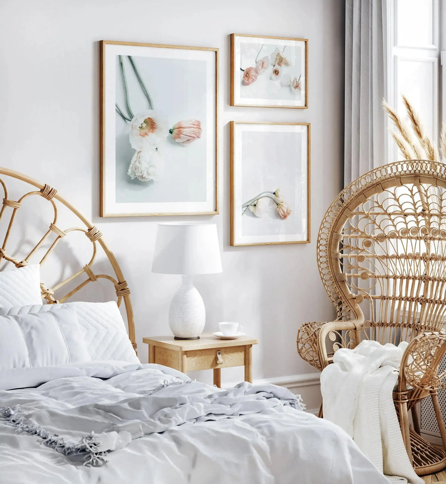 Framed art print titled Summer Poppies I displayed on a white bedroom wall in a natural wood frame with white border. The watercolor illustration features delicate pink and coral poppies with green stems arranged vertically, rendered in soft, muted tones against a pale blue-white background. The print is shown in a bedroom setting alongside a rattan peacock chair, wooden nightstand with white ceramic lamp, and natural linen bedding, creating a calm, botanical aesthetic.