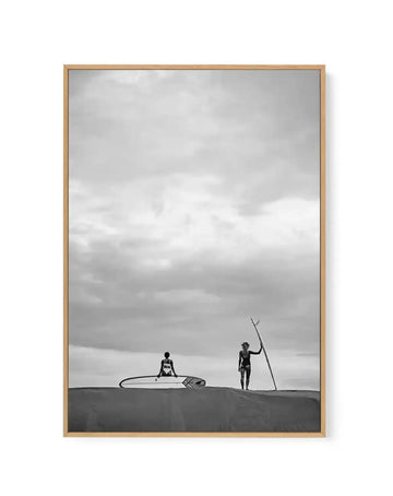 Framed black and white canvas print of a desolate beach scene with a natural wood frame. The composition features two surfers on a sandy dune under an overcast sky. One surfer sits on a surfboard, while the other stands holding a surfboard upright.
