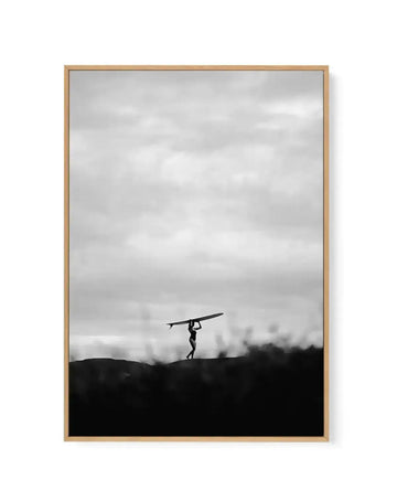 Framed black and white canvas print of a lone surfer walking along a dark, silhouetted horizon against an overcast sky. The surfer, a small figure in the center, carries a surfboard over their head, with the dark foreground suggesting a dune or hill.