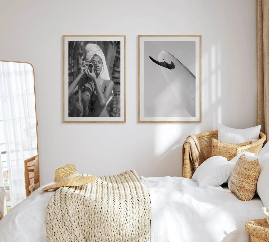 Black and white art print displayed in natural wood frame with white border, hung on white bedroom wall above woven rattan bed. Left print shows woman with head wrap holding vintage camera. Right print shows abstract curved form with black brushstroke. Bedroom styled with white linens, cream knit throw, woven pillows, and natural wood furniture. Sheer white curtains filter sunlight across scene.