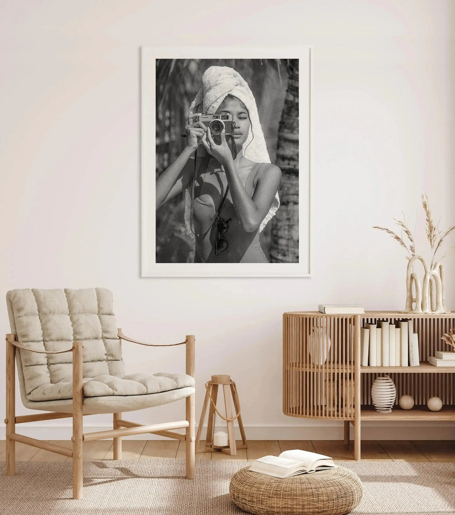 Lifestyle shot of a minimalist living room featuring a framed black and white art print of a woman with a towel on her head taking a photo with a vintage camera, with a white frame and white border. The room includes a light wood armchair with cream cushions, a small wooden side table with a candle, a woven ottoman with an open book, and a slatted wooden cabinet with books and decorative vases.