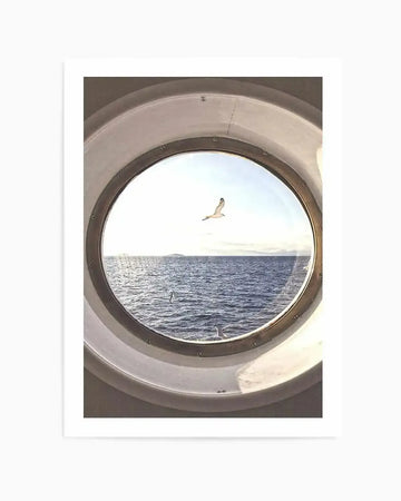 Framed photography art print of a seagull flying over the ocean, viewed through a ship's porthole. The circular composition captures the bird in mid-flight against a bright sky, with calm blue water and a distant landmass under a soft horizon. The porthole frame is a light, neutral colour with visible bolts, creating a sense of looking out from a vessel.