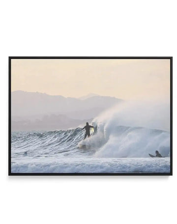 Framed canvas art print featuring a photograph of a surfer riding a large wave at dawn or dusk. The surfer in dark wetsuit stands on their board mid-wave with spray and foam visible. Another surfer floats in the foreground. Mountains in muted blue and grey tones line the hazy background under a pale cream sky. The image has a soft, misty atmospheric quality with cool tones throughout. The canvas is mounted in a black frame with white border. Horizontal landscape orientation.