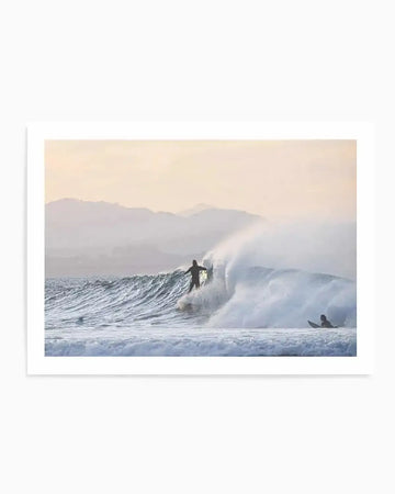 Framed photography art print of a coastal surfing scene with a natural wood frame and white border. The composition features a surfer riding a large wave, with another surfer waiting in the water, against a hazy backdrop of distant mountains and a pale orange sky.