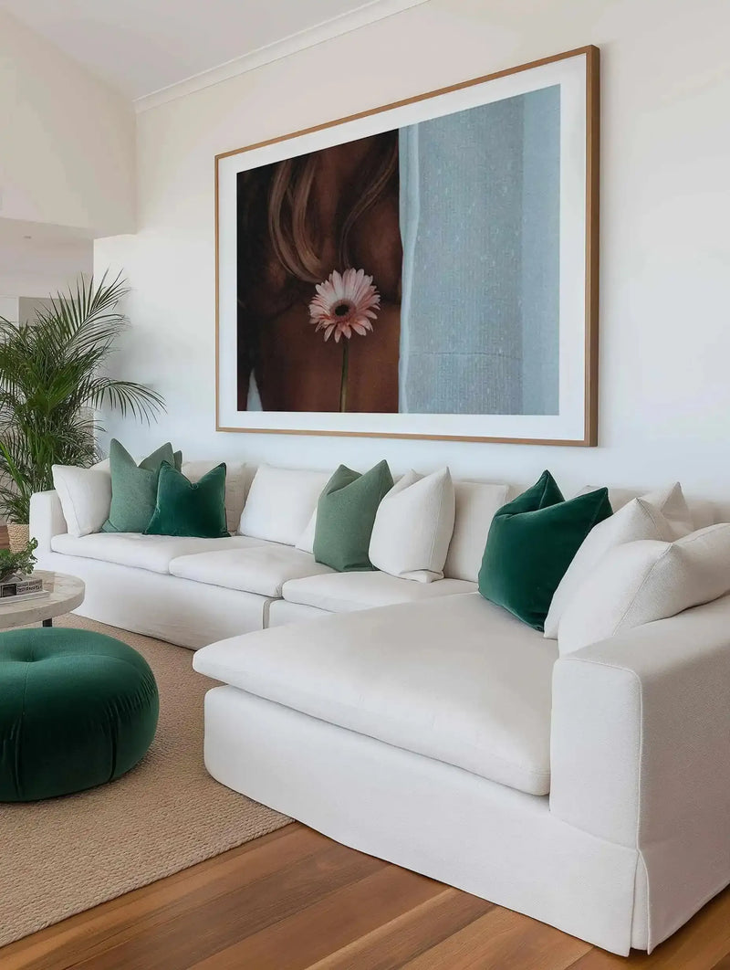 Large framed art print displayed above a white sectional sofa in a bright living room. The print is a close-up photograph of a woman's back with long, light brown hair, holding a single pink gerbera daisy with a dark center. The background is a light blue textured wall. The art print has a natural wood frame with a white border. The sofa is adorned with green and white throw pillows, and a round green velvet ottoman sits on a natural fiber rug on a wooden floor.