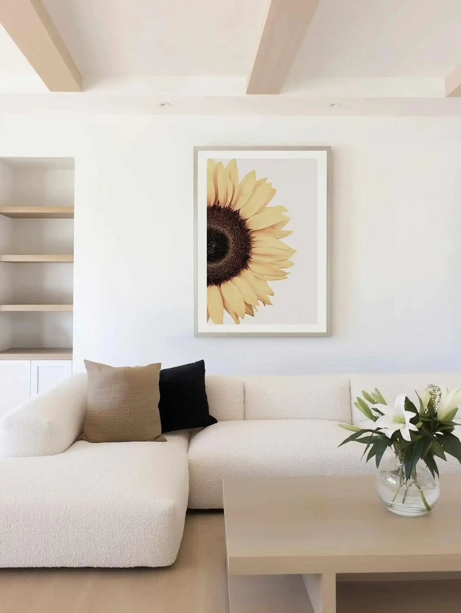 Large framed art print displayed above a curved white sofa in a minimalist living room. The print features a close-up, cropped view of a sunflower with bright yellow petals and a dark brown seed head, with a natural wood frame and white border. The room includes a light wood coffee table with a vase of white lilies.