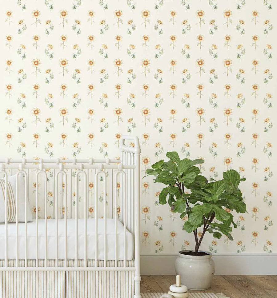 Lifestyle shot of a nursery with a white crib and a potted plant, featuring peel and stick wallpaper with a repeating pattern of hand-painted sunflowers in warm yellow and orange with green leaves on a soft cream background. The botanical design has a loose, organic watercolour quality.