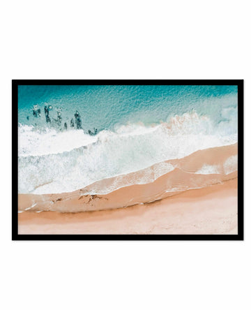 Aerial photograph framed in black showing a coastal beach scene with turquoise ocean water, white foaming waves, and sandy beach in warm tan and peachy tones. The composition captures multiple wave layers rolling toward shore with a group of people visible as small figures in the shallow turquoise water on the left side. The image has a serene, minimalist quality with soft, natural colors transitioning from deep teal at the top to pale sand in the foreground.