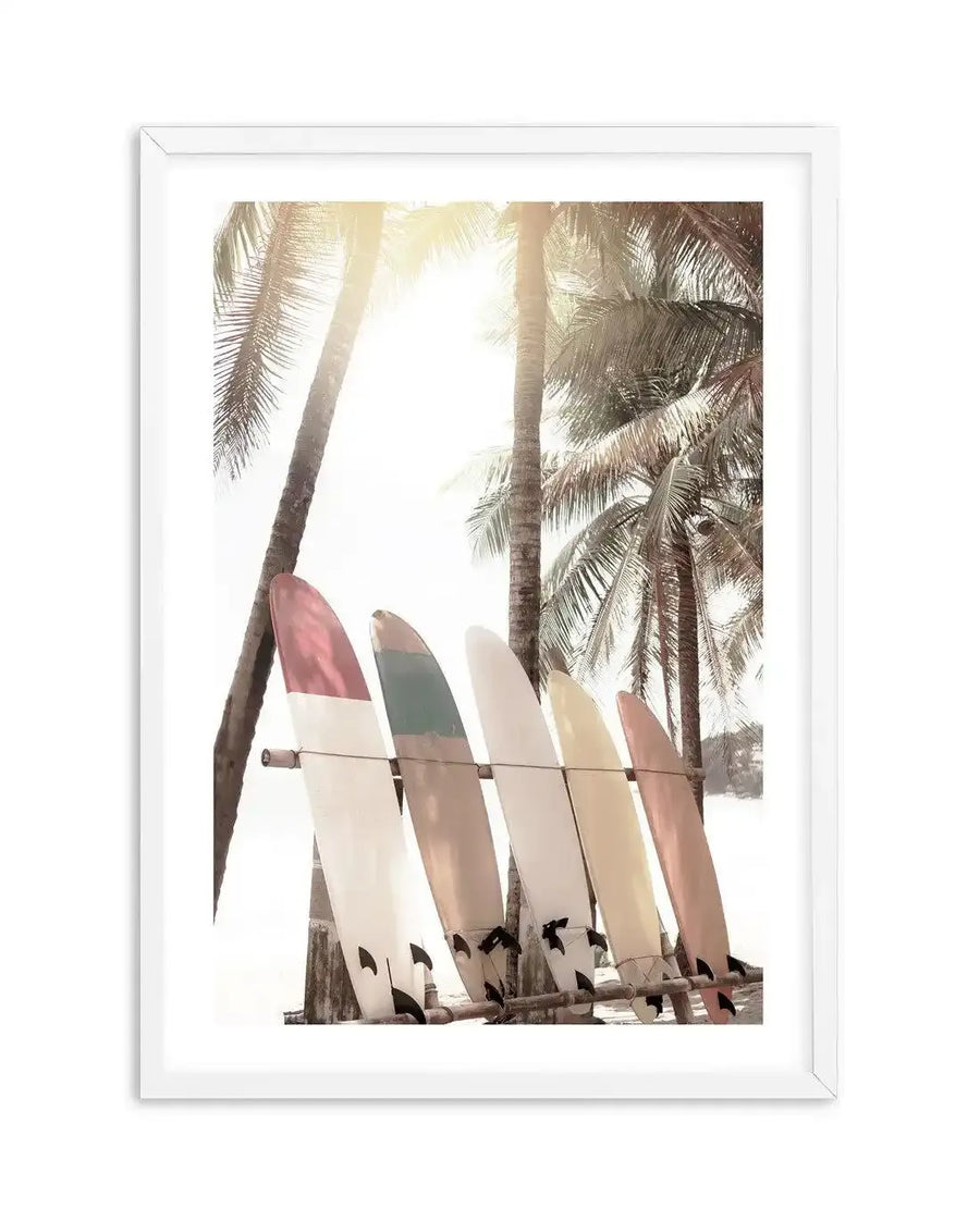 Framed photography art print of a tropical beach scene with a bright, sun-drenched sky and tall palm trees. Five surfboards in shades of red, green, cream, yellow, and pink are propped against a wooden rack in the foreground, with a natural wood frame and white border.