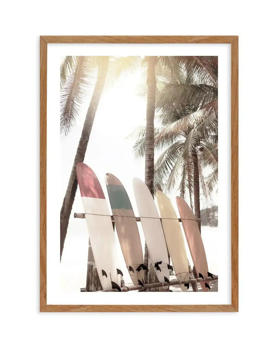 Framed photography art print of a tropical beach scene with five surfboards leaning against a wooden rack, displayed in a natural oak frame with white border. The composition features tall palm trees with bright sunlight filtering through the fronds, creating a warm, hazy glow. The surfboards are in various pastel shades including red and white, green and white, cream, yellow, and pale pink.