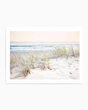 Framed photography art print of a serene beach scene with a soft, diffused light. The foreground features white sand dunes with sparse tufts of light green and golden beach grass. In the midground, gentle waves with white foam break on the shore, and the background shows a hazy, light blue ocean meeting a pale, almost white sky. The overall mood is peaceful and airy.