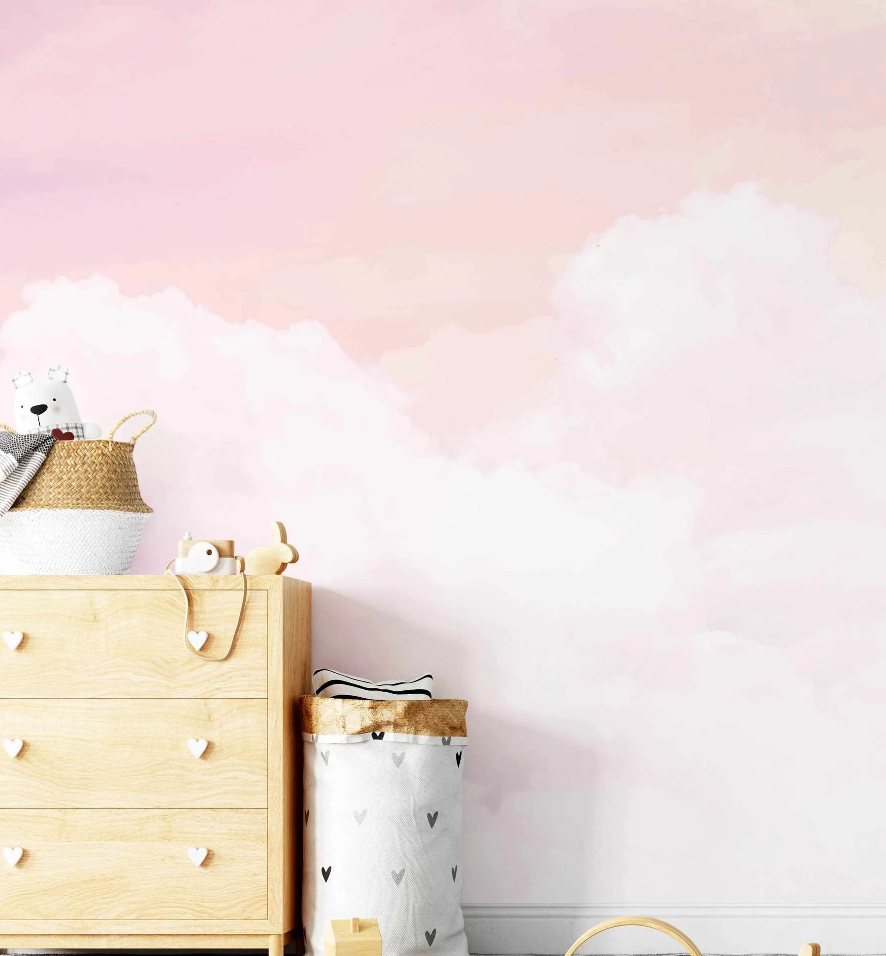 Lifestyle shot of a child's room featuring a peel and stick wallpaper mural with a soft, dreamy watercolor design of pink and white clouds against a pale pink sky. The wallpaper covers the entire wall behind a light wood dresser with heart-shaped knobs, a woven basket with a white teddy bear, and a white and grey heart-patterned laundry bag.