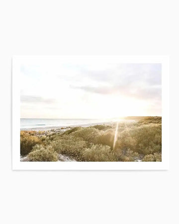 Framed photograph of a coastal beach scene at sunset with a natural wood frame and white border. The composition features a sandy beach and ocean under a bright, hazy sky, with the sun low on the horizon casting a strong lens flare over a foreground of golden-hued coastal shrubs.