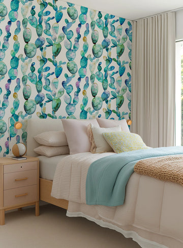 Lifestyle shot of a bedroom featuring a repeating pattern of hand-painted prickly pear cacti in various shades of blue, green, and purple on a white background. The wallpaper covers the wall behind a bed with a light-colored headboard, cream bedding, and a light blue throw blanket. A light wood nightstand with a lamp sits to the left of the bed, and a window with cream curtains is on the right.