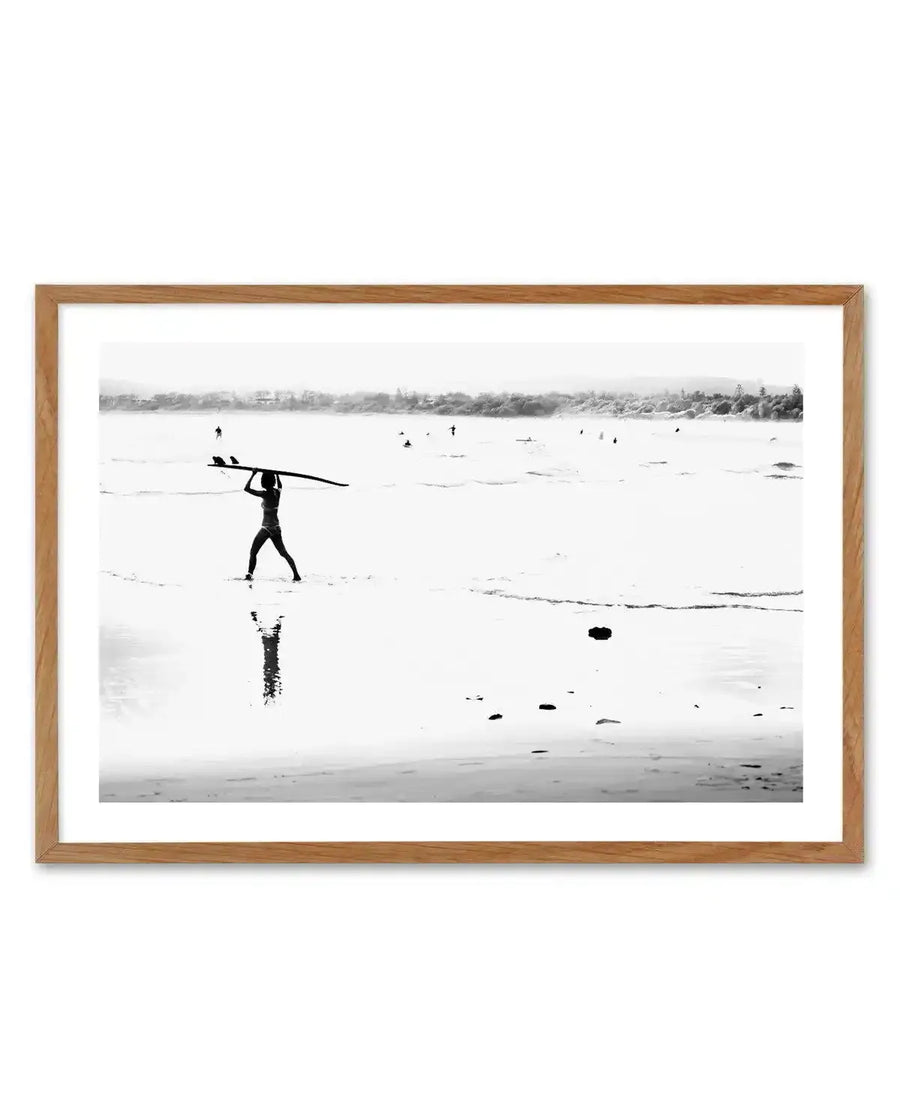 Framed black and white photograph of a coastal beach scene with natural wood frame and white border. The composition features a lone surfer walking along wet sand at low tide, carrying a surfboard overhead, with gentle waves and an overcast sky stretching to the horizon.