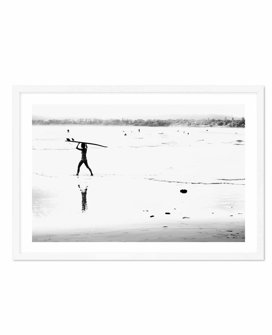 Black and white photograph framed in white, depicting a surfer in a wetsuit carrying a surfboard overhead while walking through shallow water toward the ocean. Additional surfers can be seen in the distance in the water. The composition captures a misty, overcast coastal scene with a tree-lined shoreline visible across the bay. Rocks dot the wet sand in the foreground. The photograph has a grainy, documentary-style quality with high contrast between the dark figures and the pale, hazy sky and water.