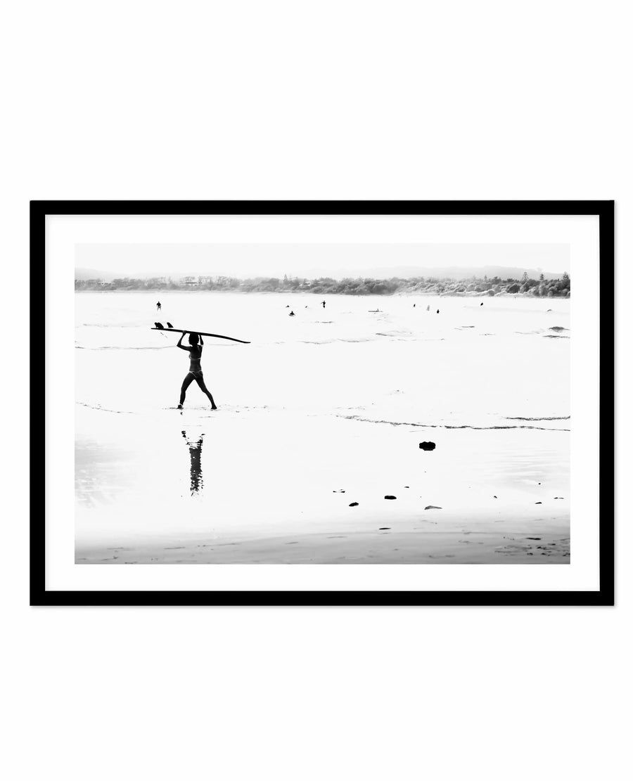 Black and white framed art print showing a surfer in a wetsuit carrying a surfboard over their shoulder, wading through shallow water. The surfer is reflected in the wet sand below. In the background, calm ocean waters with several other surfers visible in the distance, and a tree-lined shoreline. The photograph captures a serene, minimalist beach scene with soft, muted tones. Black frame with white border.