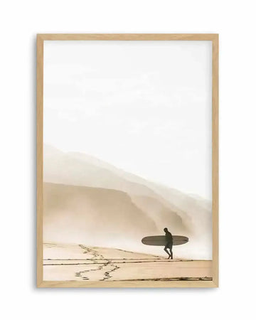 Framed photography art print of a coastal beach scene with a natural wood frame and white border. The composition features a lone surfer walking along wet sand at low tide, carrying a surfboard, with layered misty mountains in the background and an overcast sky.